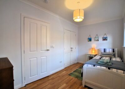 White, single bed on wooden floor. Two doors opposite with window above bed.