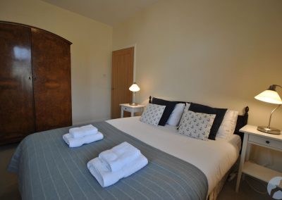 Double bed between white bedside tables with lamps. To the left of the bedhead is a door and a large wooden wardrobe.