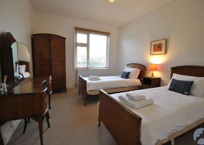 View down bedroom across two beds towards a window. To the left of the window is dark wooden wardrobe and a dressing table with mirror.