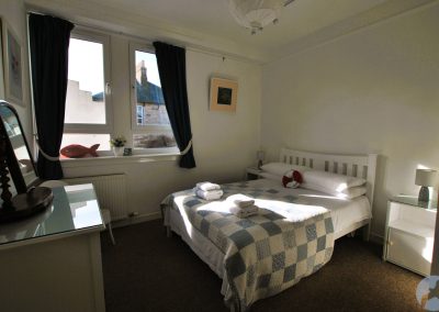 White, wooden bed frame, large window and chest of drawers.