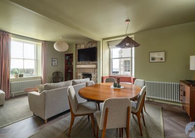 There is a fireplace with a TV above on an exposed wall between two windows. In the foreground there is a round dining table woth six chairs.