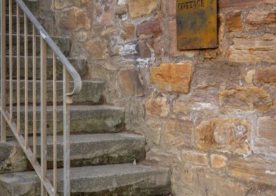Stone steps with a metal railing leading up towards the front door. A sign on the stone wall reads Ledger's Cottage.