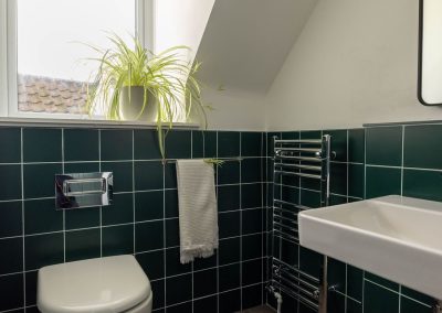 Corner of the shower room beneath a sloped ceiling. There is a window above a tiled green wall up to waist level.