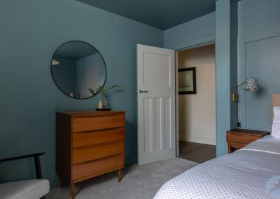 A door lies open from the bedroom into a hallway. There is a circular mirror above a chest of drawers.