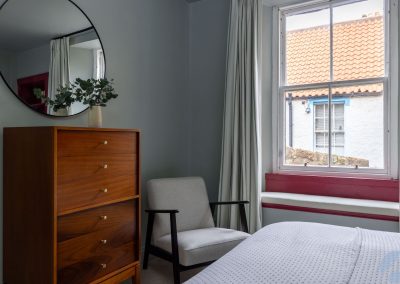 A circular mirror hangs above a chest of drawers to the left of an armchair and window.