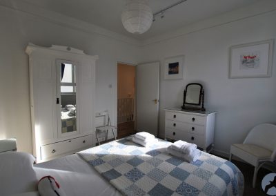 View from headboard across a blue and white checkered bedspread towards the door with wardrobe and chest of drawers either side.