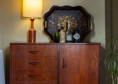 A lamp, tray and bottles of gin sit on top of a vintage-style sideboard with drawers.