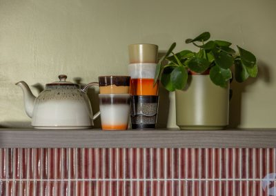 1970s style kettle and mugs on a shelf above a deep red tiled wall.