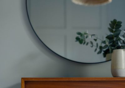 Reflection of a pendant light in a round mirror above a chest of drawers.