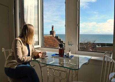 Woman seated at table looking out of an open window towards the sea.