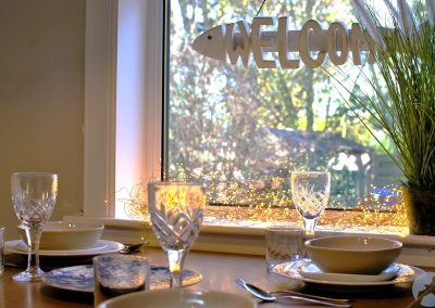 Dining table set with plates, glasses and cutlery beside a window, styled with soft lighting, greenery and a decorative welcome sign.