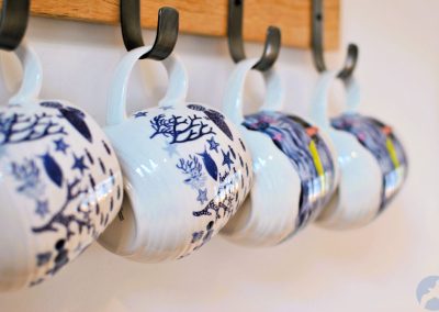 Row of patterned ceramic mugs hanging on hooks in the kitchen, featuring coastal-inspired designs in blue and white.