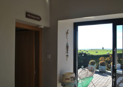 Interior view showing a doorway with a “Fife Coastal Path” sign and a glazed door opening onto a deck with seating and open countryside views beyond.