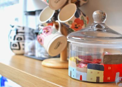 Kitchen worktop with hanging patterned mugs and a glass jar with a painted seaside village design holding tea and coffee sachets.
