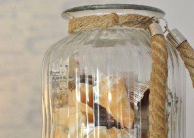 Glass jar filled with seashells, wrapped with a natural rope handle, displayed on a white surface in soft light.