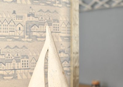 White wooden sailboat ornament on a shelf, set against patterned wallpaper with coastal village illustrations.