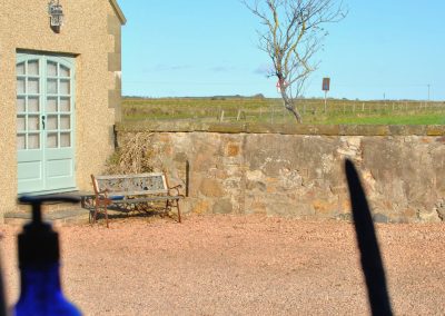 View from inside the cottage looking out across a gravel driveway to a stone wall, bench and open farmland beyond under a clear blue sky.
